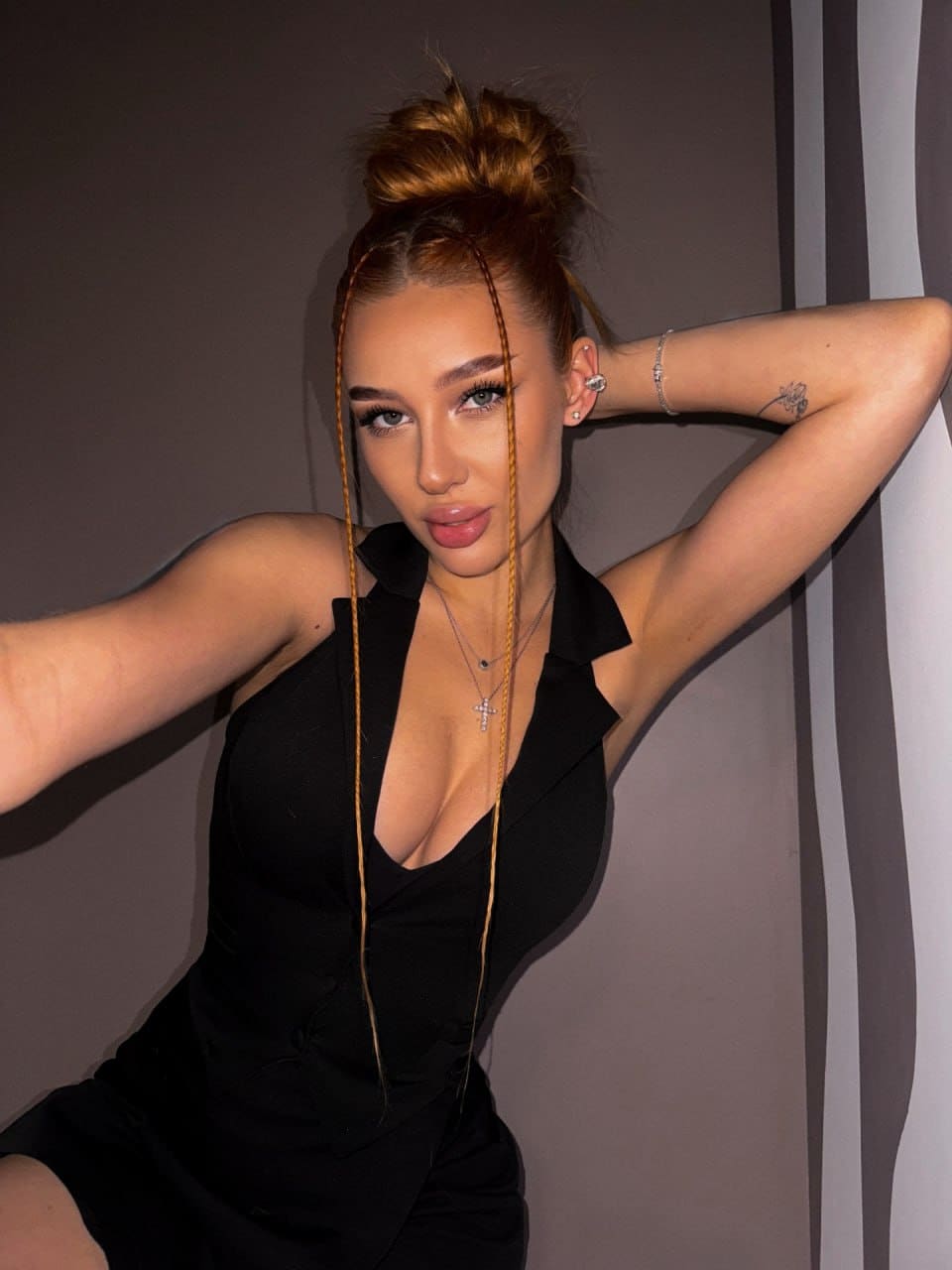 Against a striped wall, she poses with an arm raised, wearing a black plunging top, cross necklace, and bracelet.