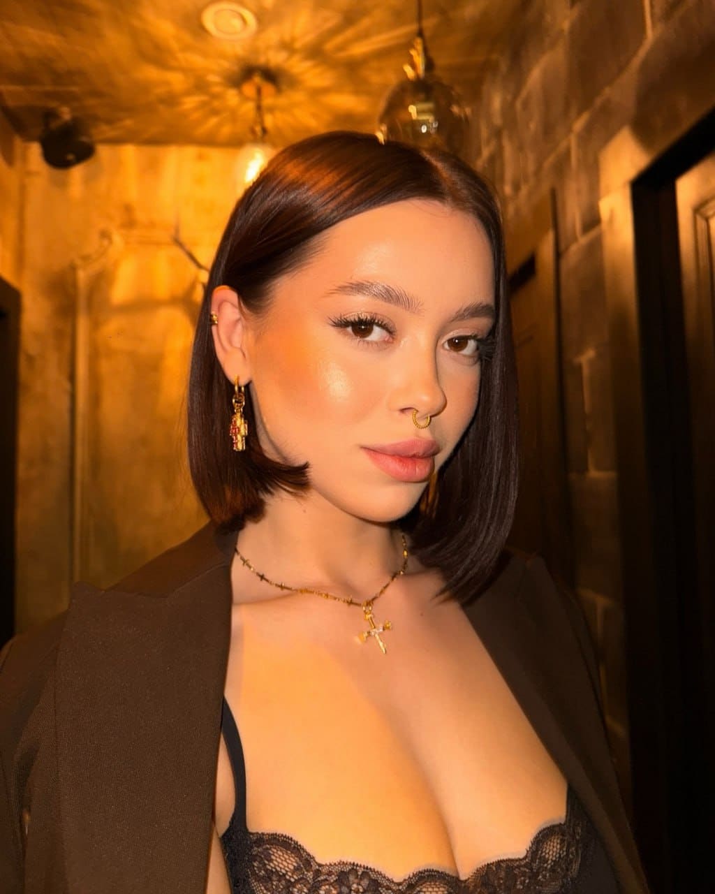 In a dim hallway, she wears a black lace bra under an open dark blazer, accessorized with a gold cross necklace and dangling earrings.