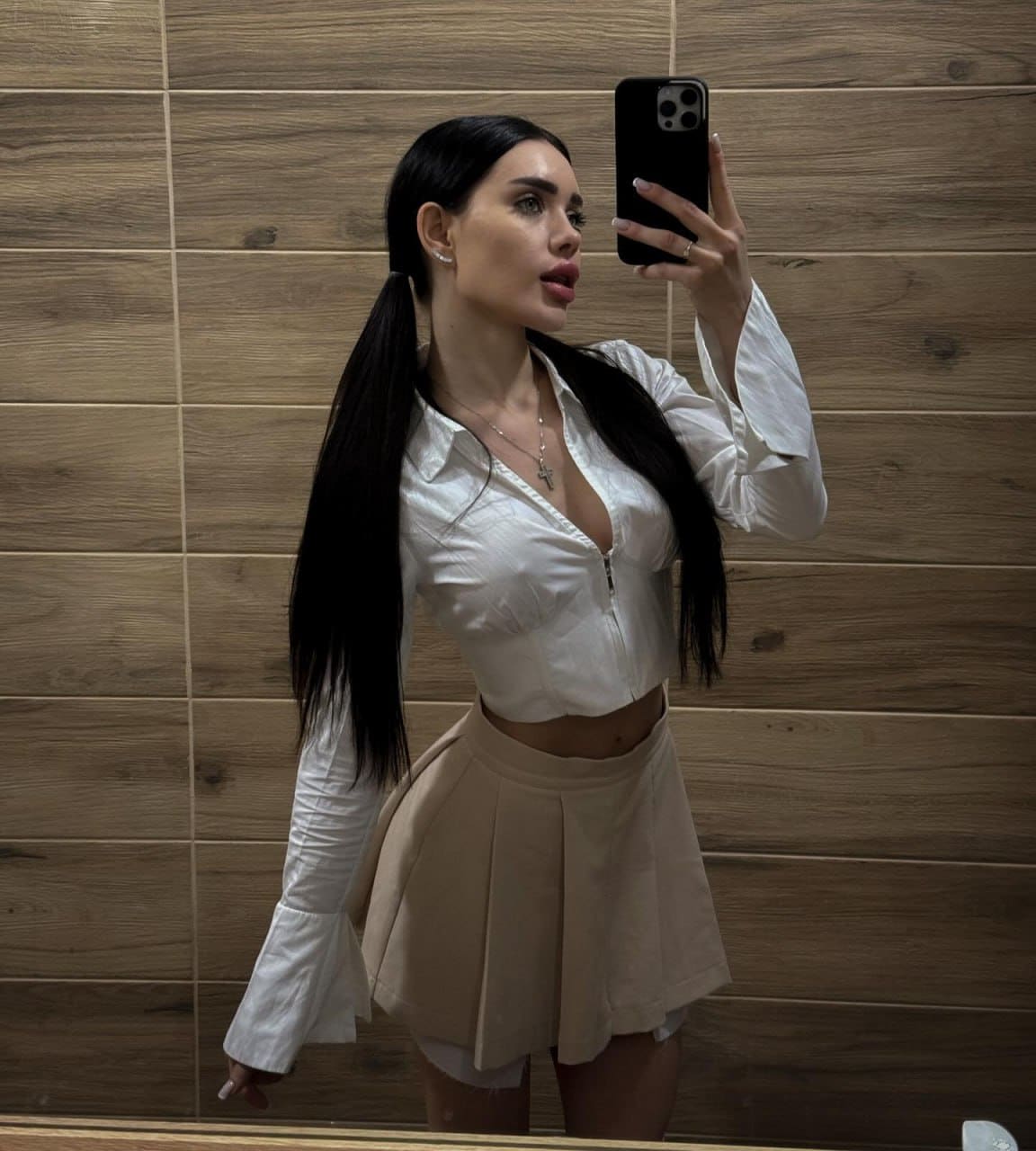 A mirror selfie shows Karolina in pigtails, wearing a white cropped zip-up and beige pleated mini skirt against a wood-tiled wall.