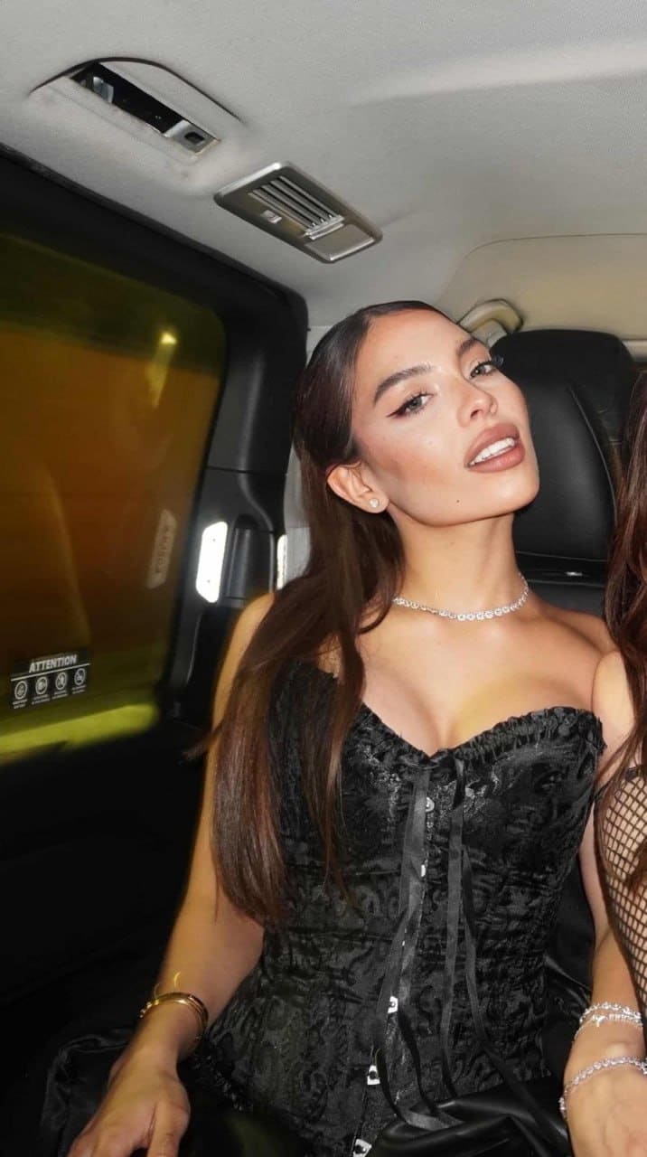 Seated inside a vehicle, Alana wears a black lace-up corset top and silver necklace, her long dark hair pulled back in this close-up portrait.