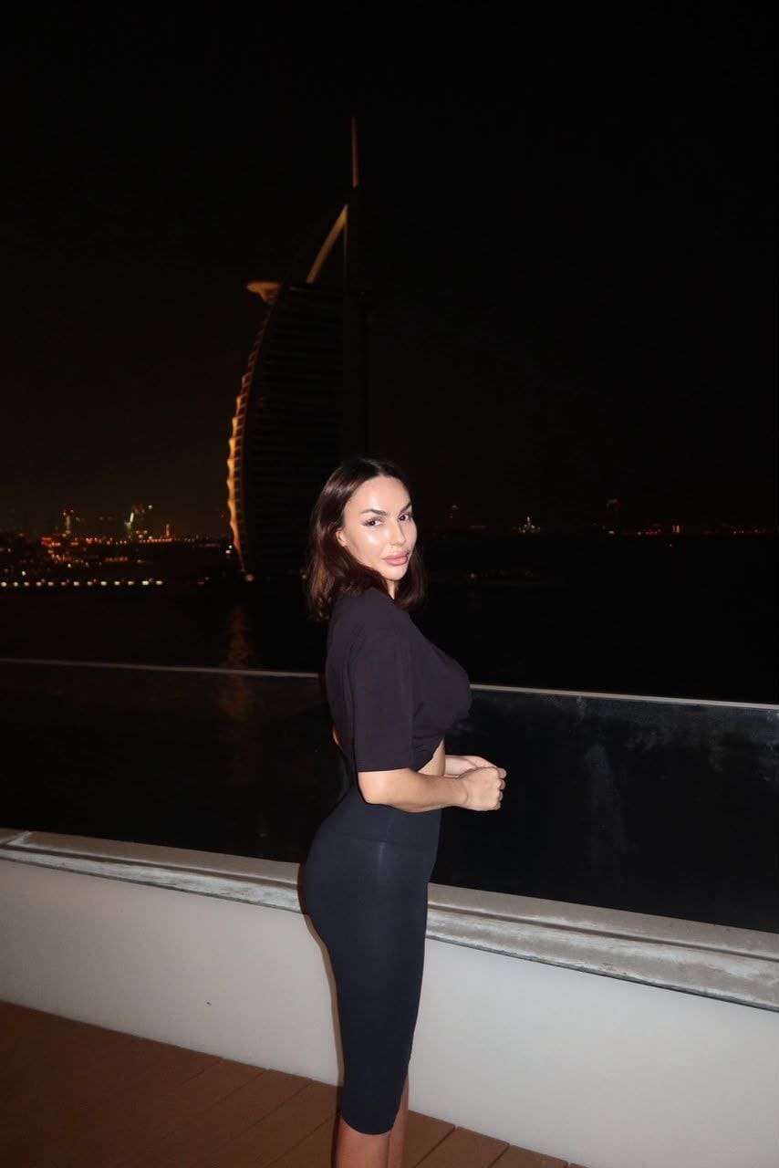 Standing in profile on a night balcony, she wears a black cutout midi dress with the sail-shaped skyscraper in the background.