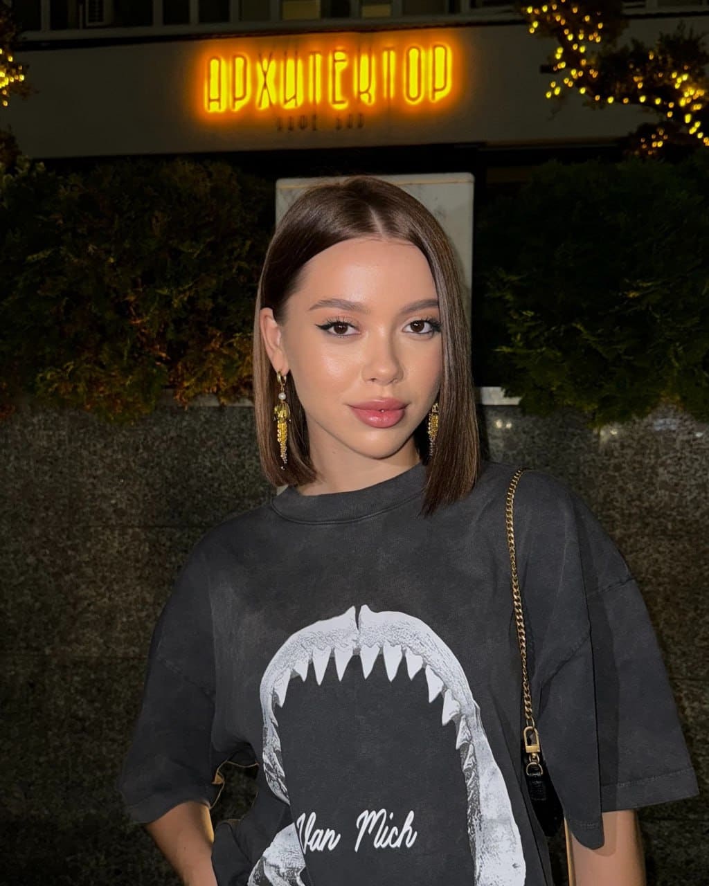 Outdoors at night near a lit sign, she wears an oversized black t-shirt with a shark mouth graphic and a chain-strap bag.