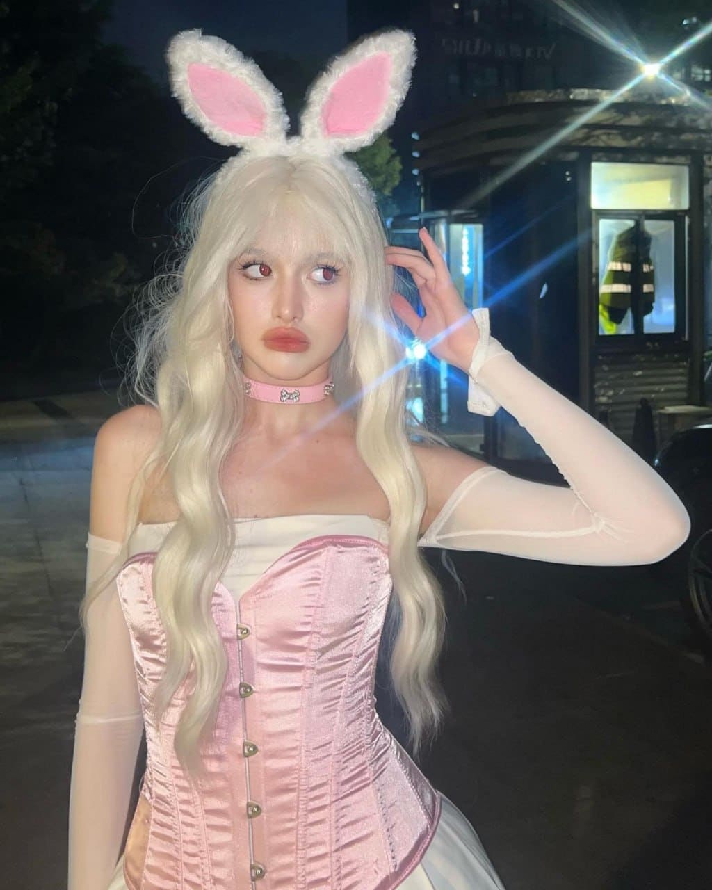 At night, a person with platinum blonde hair and bunny ears wears a pink satin corset with sheer sleeves.
