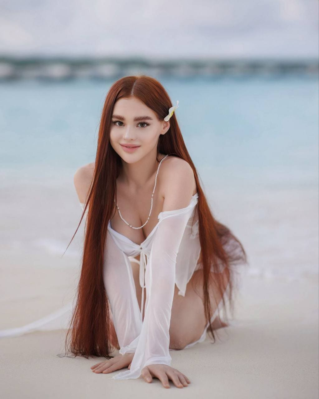 Kneeling on sand, Georgia wears a sheer white off-the-shoulder cover-up over a bikini, with a white flower tucked behind her ear.