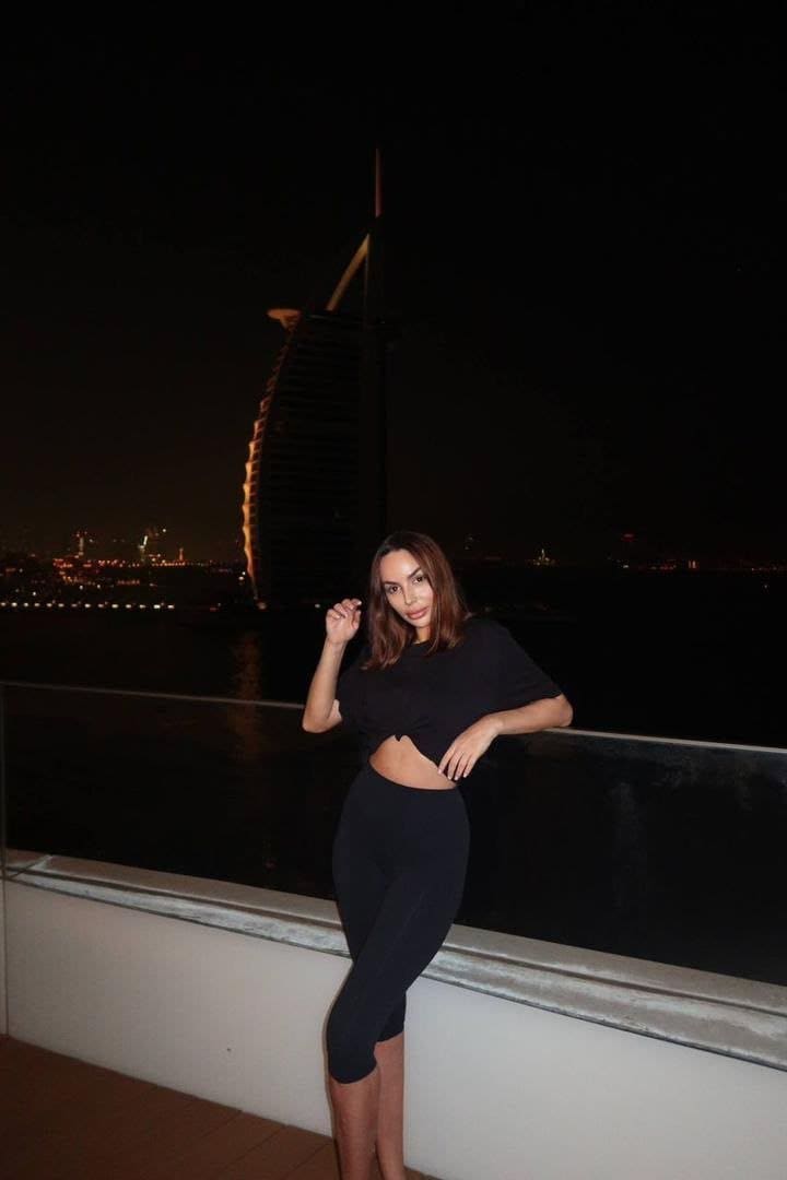 Leaning on a balcony railing at night, Nicole wears a black crop top and leggings with a lit sail-shaped building behind.