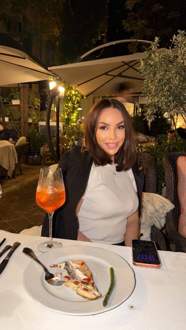 At a night dining table, she wears a white ribbed top and black blazer, with an orange drink and food in the foreground.