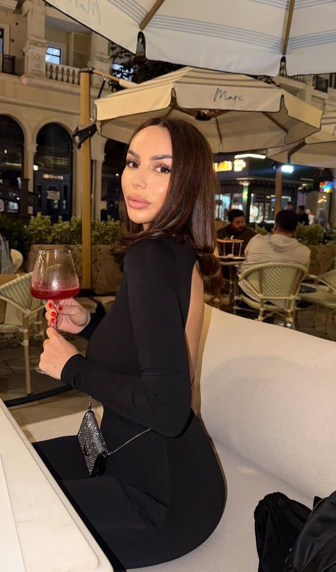 Seated in profile at an outdoor table, she holds red wine in a long-sleeved black dress with an open back.