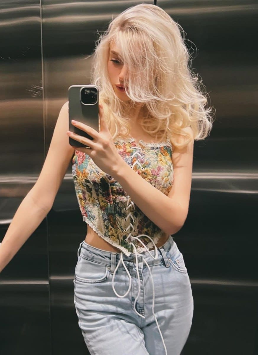 Taking a mirror selfie in an elevator, Laura wears a floral lace-up corset top with spaghetti straps and light-wash blue jeans.