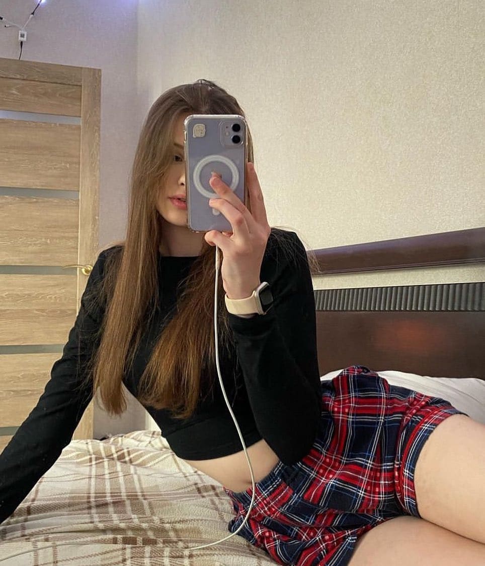 Sitting on a bed, Ava takes a mirror selfie wearing a black long-sleeved crop top and a red plaid mini skirt that exposes her midriff and legs.