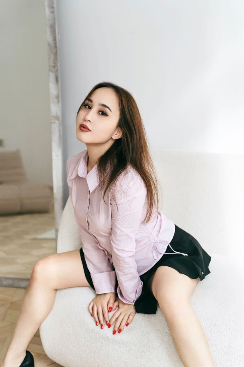 Sonya sits on a white chair in a pink shirt and black skirt, legs spread with hands between thighs.