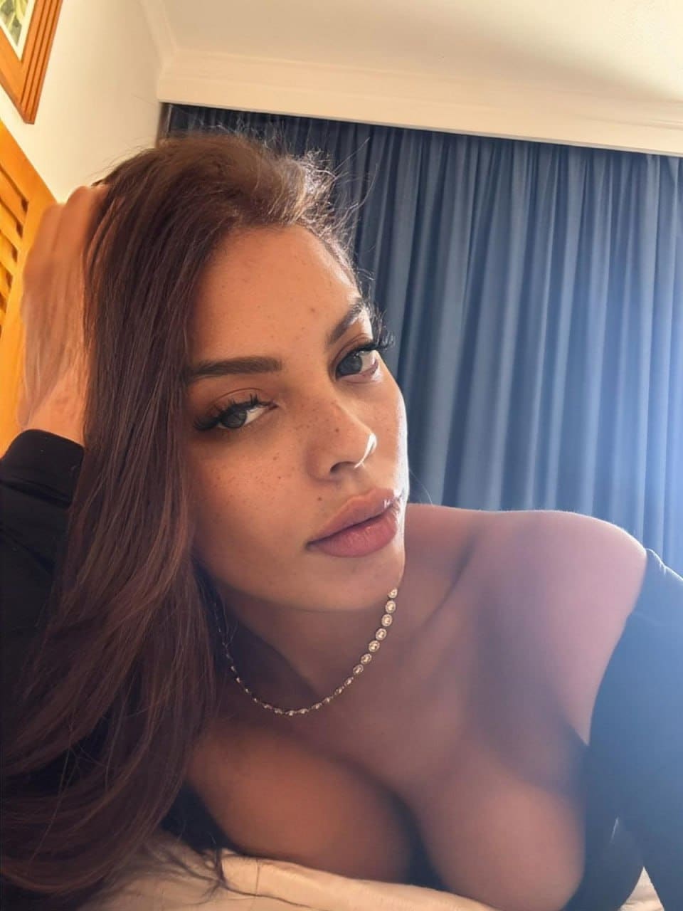 Reclining on a bed against blue curtains, Alana runs a hand through her brown hair while wearing a black off-the-shoulder top.