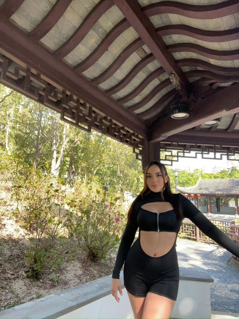 Under a wooden pergola, she wears a black long-sleeved bodysuit with a high neck, chest cutout, zipper, and midriff cutout, arm extended.