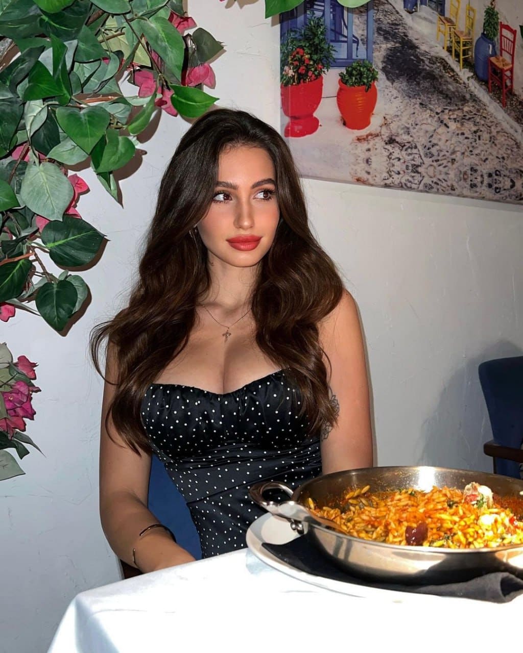Sophia sits at a table with a pan of food, wearing a strapless black dress with white polka dots and a sweetheart neckline against a white wall.