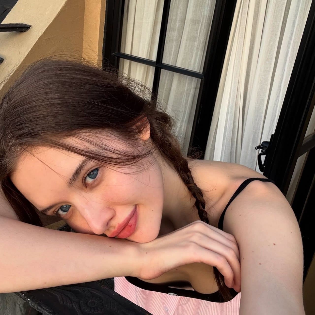 Resting her chin on crossed arms, she wears a pink and white striped top against sheer white curtains.