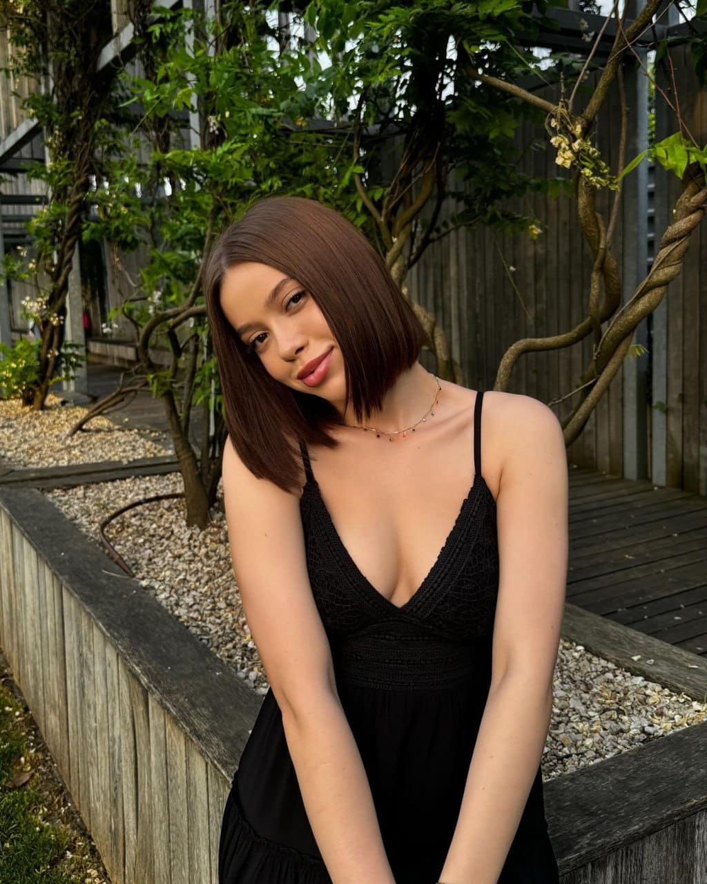 Juliana tilts her head in a black spaghetti-strap dress with a deep V-neck, standing outdoors near a wooden planter and climbing vines.