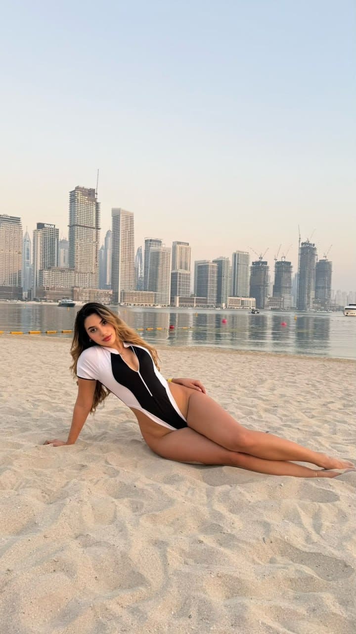 Reclining on a sandy beach with a city skyline behind, Sammy wears a black and white one-piece swimsuit with short sleeves.