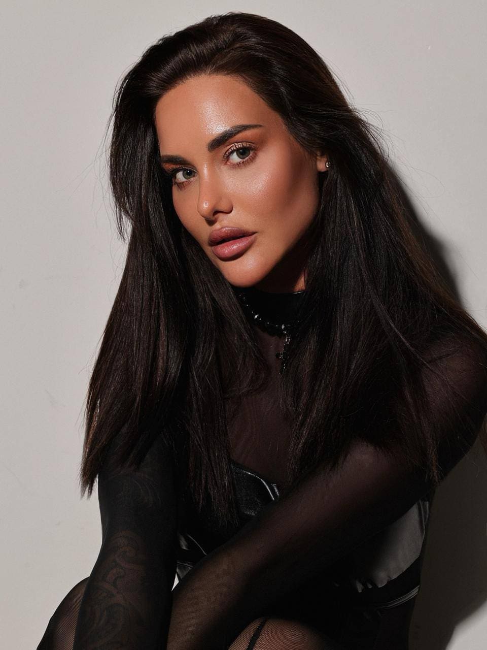 Eliana poses with arms crossed, wearing a sheer black mesh top over a dark undergarment against a plain background.
