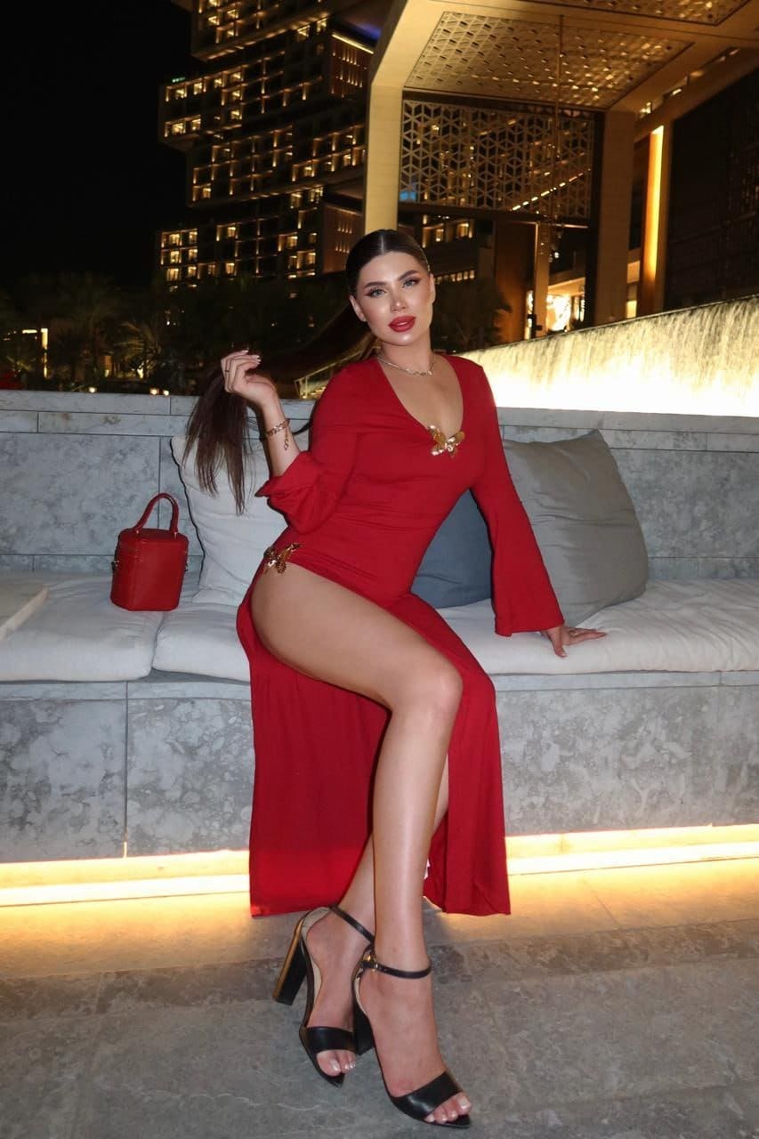 Seated on an outdoor sofa at night, Lena wears a red long-sleeved dress with a plunging neckline and high slit, paired with black strappy high heels.