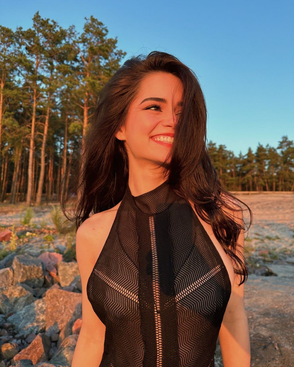 Juliana stands smiling in a black halter-neck top with a zipper, set against a pine forest at dusk.