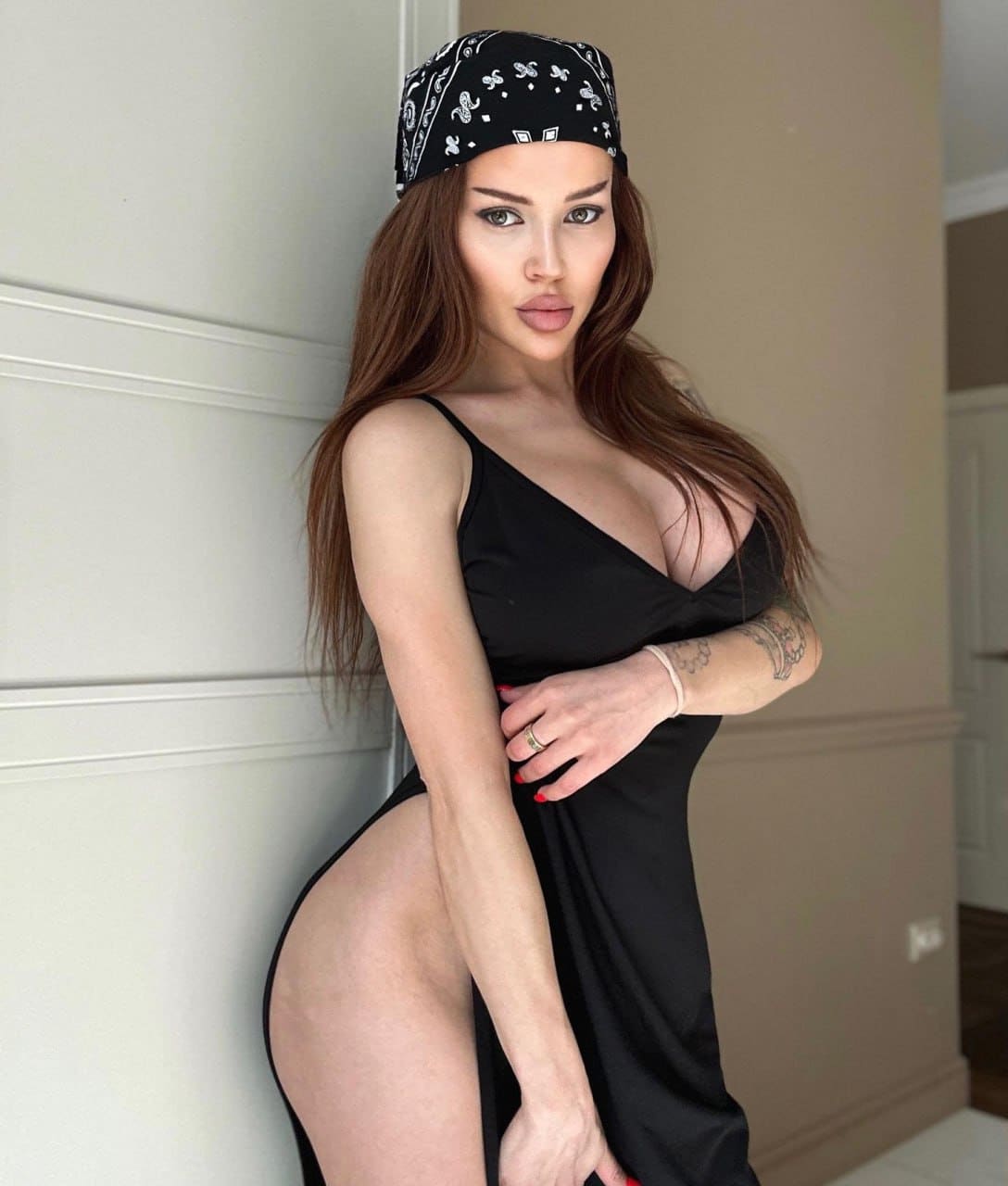 Against a white paneled wall, she stands in a three-quarter pose wearing a black paisley bandana and a satin slip dress with a deep V-neck and thigh slit.