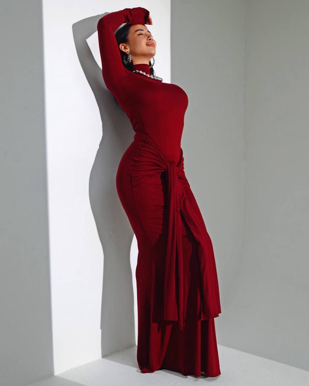 Malvina stands in profile against a white wall, arms raised above her head, wearing a floor-length burgundy dress with a high neck.