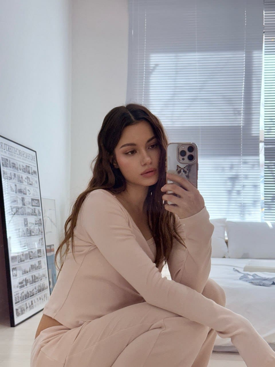 Mira sits on the floor taking a mirror selfie in a matching light pink long-sleeved top and sweatpants, with a bed and framed poster behind her.