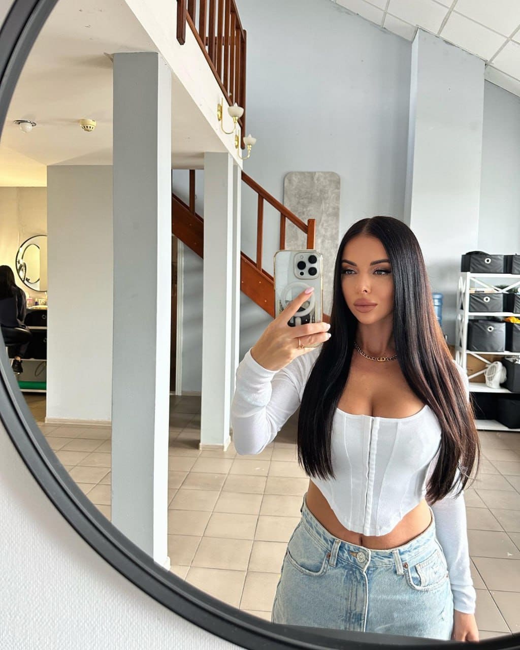 A mirror selfie shows her in a white corset top and denim skirt, standing near a wooden staircase indoors.