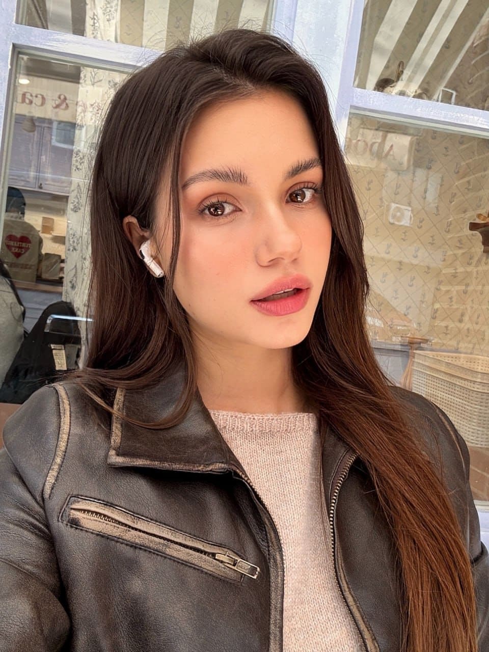 Mira takes a selfie in front of a windowed storefront, wearing a dark leather biker jacket over a light pink knit sweater with a white earbud.