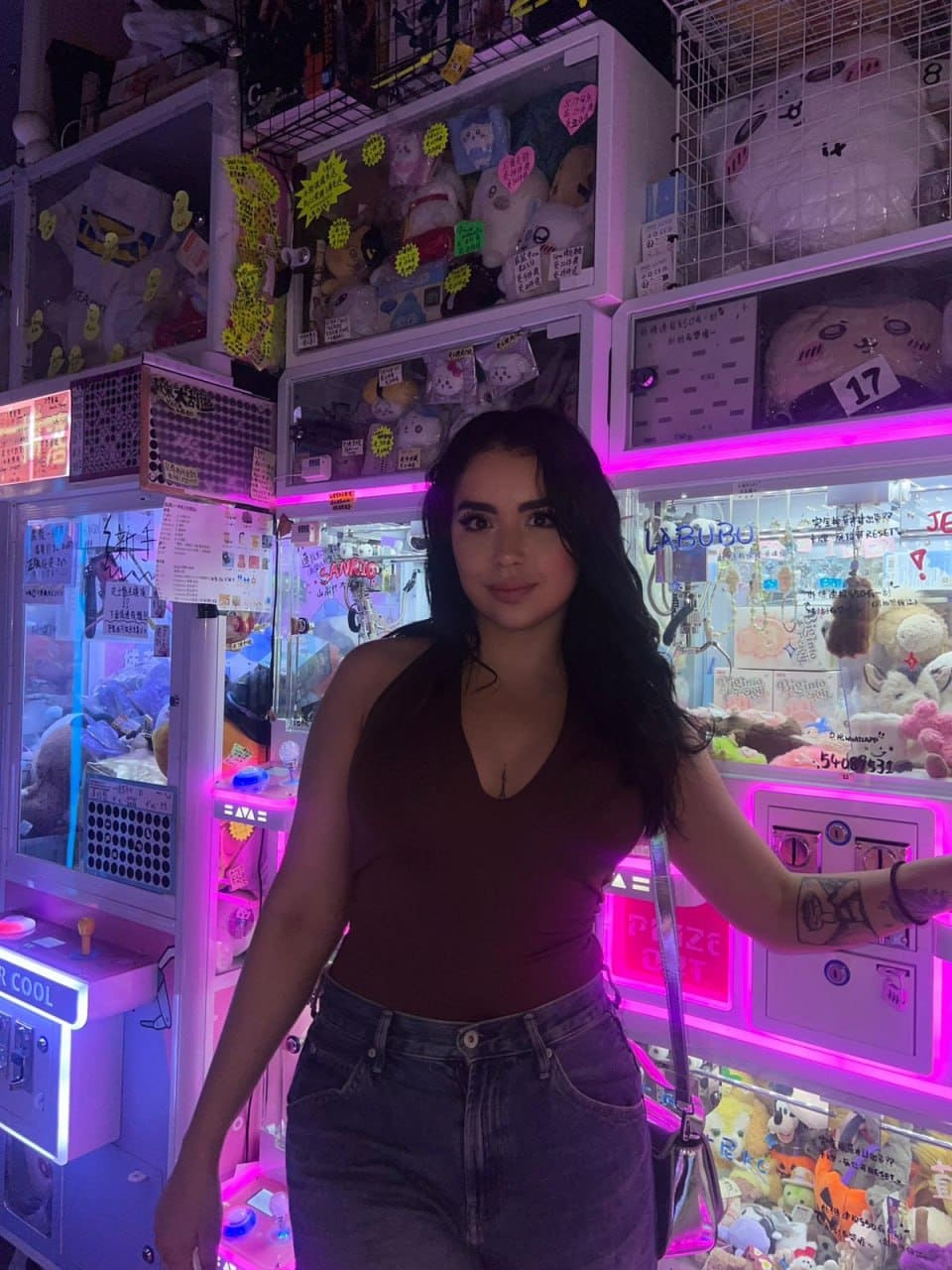 Rosa stands before illuminated claw machines in a dark maroon sleeveless top with a deep V-neck and blue jeans, arm tattoo visible.