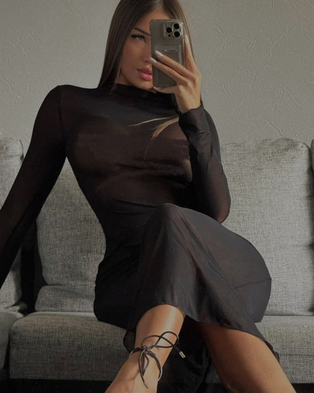Seated on a gray sofa for a mirror selfie, she wears a sheer black midi dress and strappy heels, revealing her silhouette.