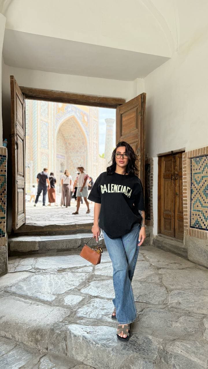 Standing on stone steps in a tiled entryway, a woman with glasses wears an oversized black t-shirt, blue jeans, sandals, and holds a brown bag.