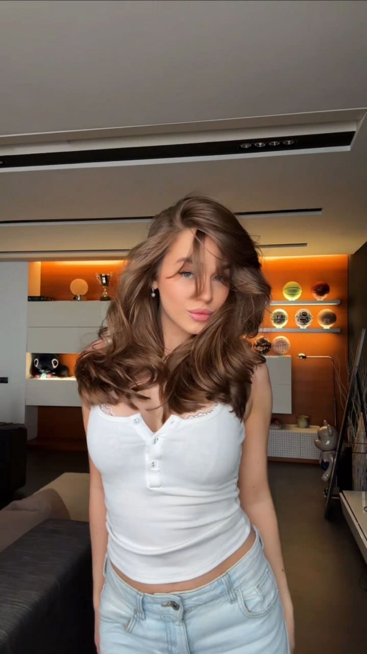 She poses indoors with a tilted head in a white buttoned henley and light blue jeans, set against warm orange lighting and background shelving.