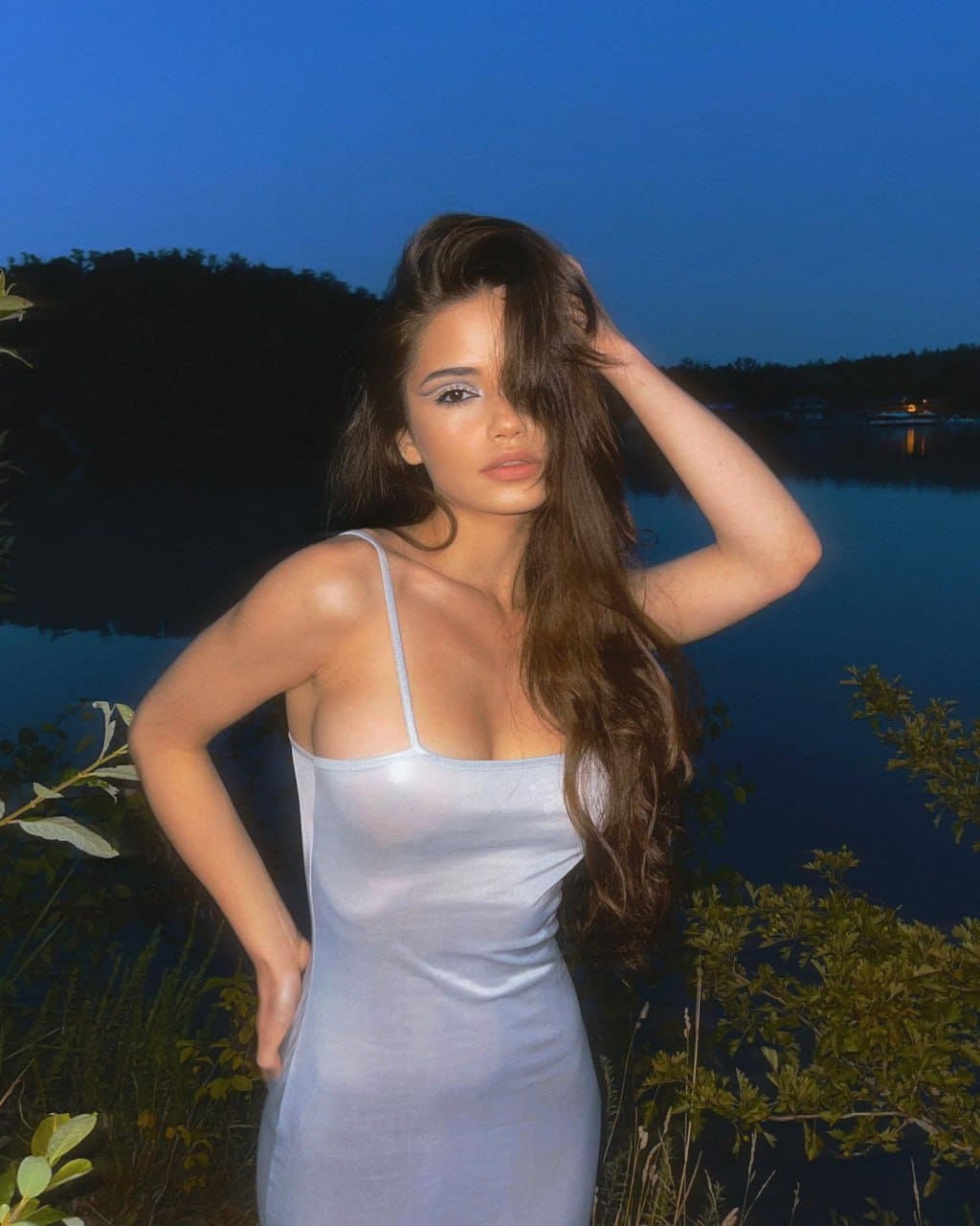 By water at dusk, Juliana poses in a light sleeveless dress, one hand raised to her hair.