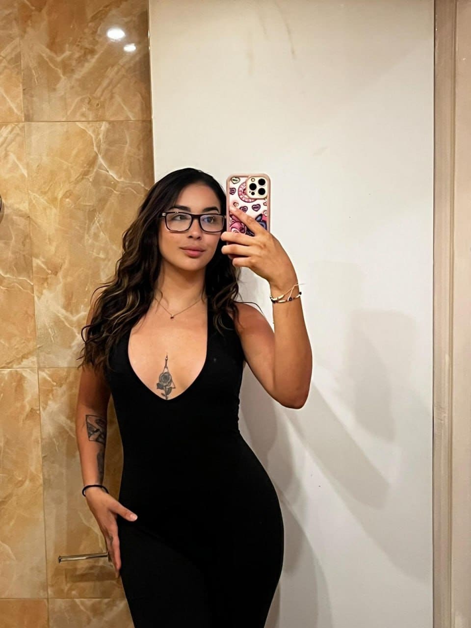 In a mirror selfie, Rosa wears glasses and a black sleeveless bodysuit with a deep V-neck, revealing a chest tattoo in a marble room.