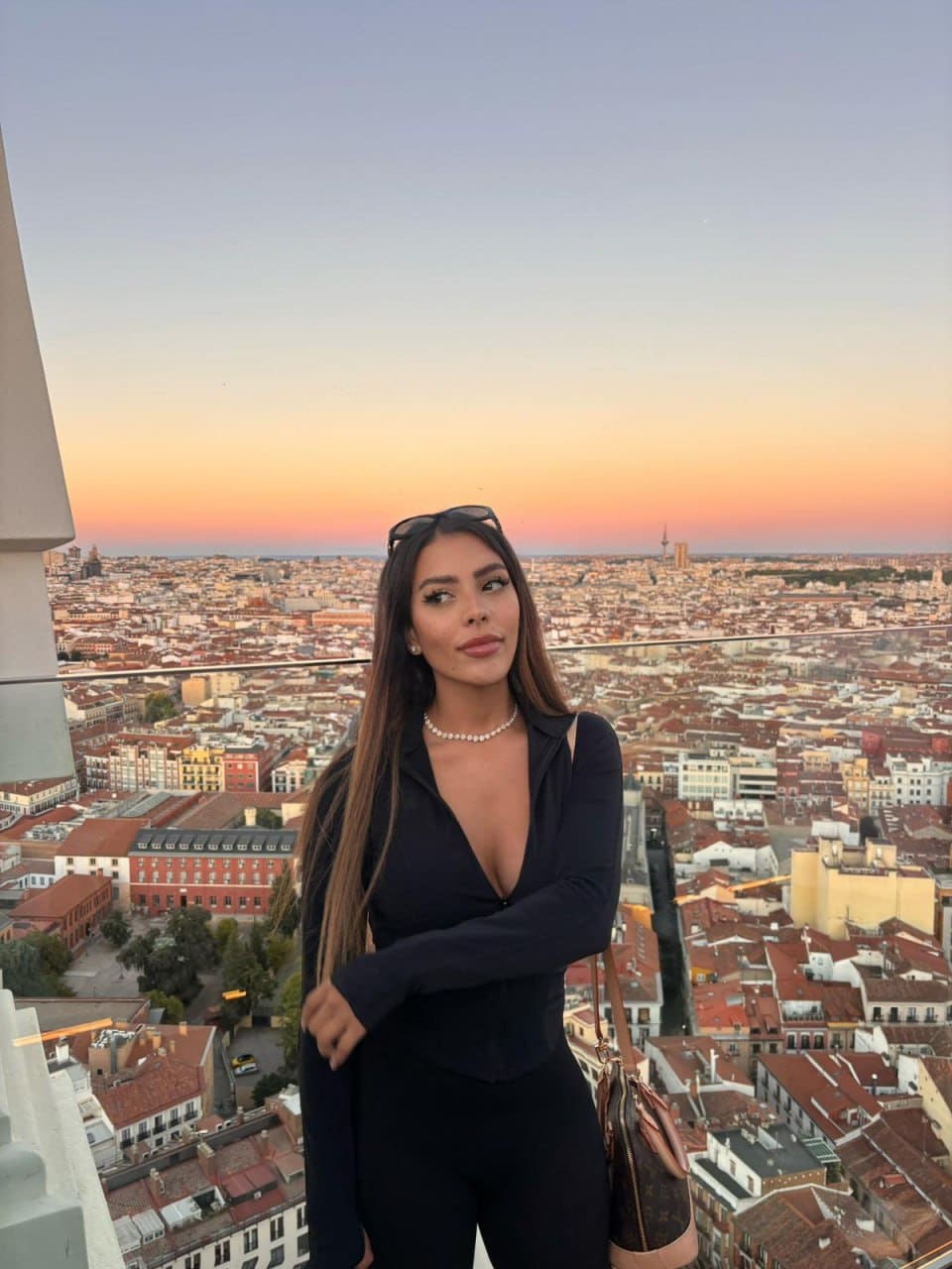 Alana stands on a rooftop terrace at sunset in a black plunging bodysuit, pearl necklace, and sunglasses, holding a handbag against a city skyline.