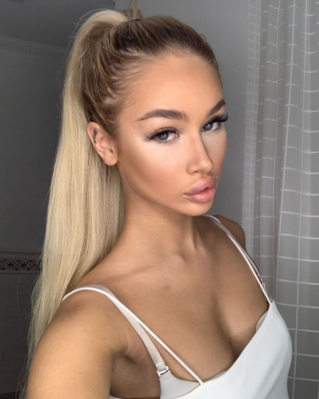 Stella poses indoors against a grey wall, wearing a white top with thin double straps and her blonde hair in a high ponytail with side braids.